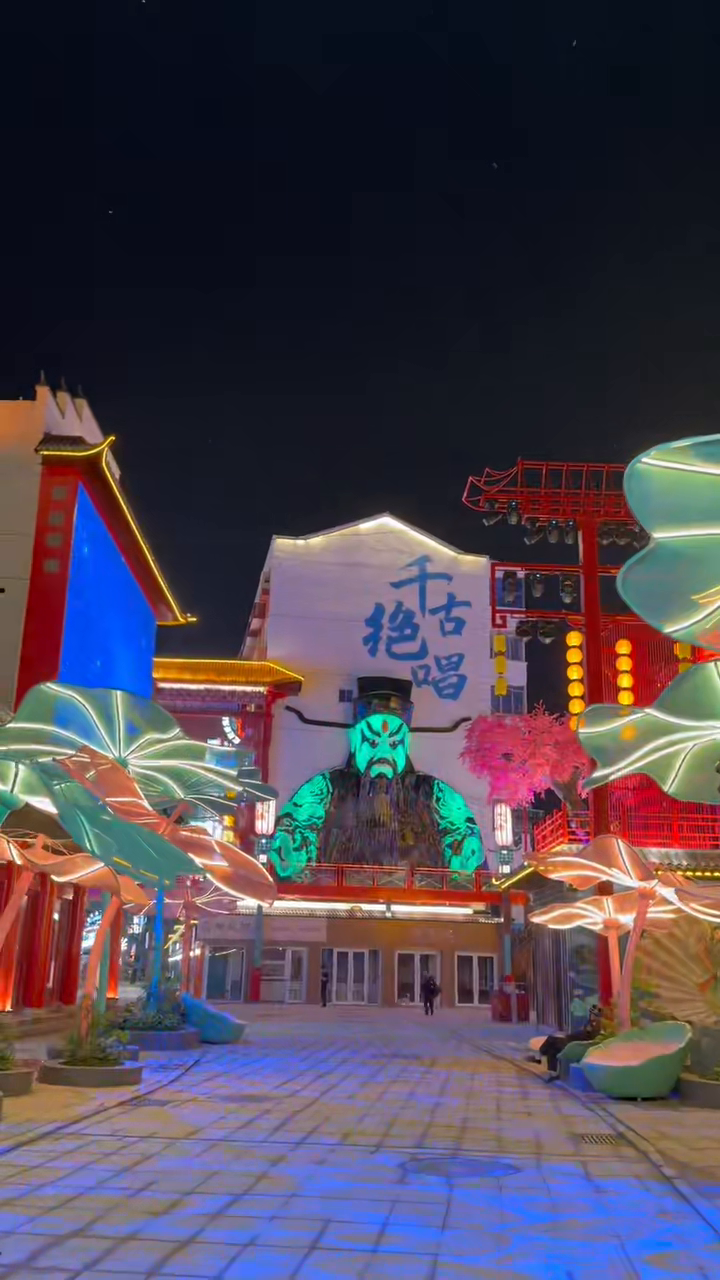 Green-lit Chinese opera-mask figure between illuminated leaf-shaped elements in a night courtyard.